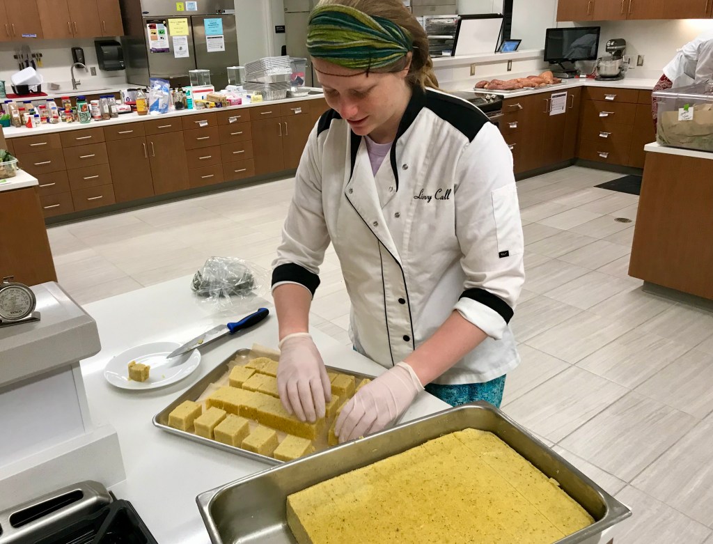 Person with a personalized chefs coat arranging cubes of yellow food on a sheet tray in a large kitchen.