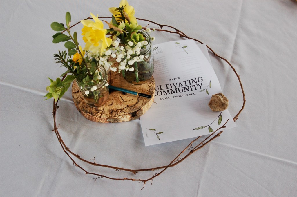 A round tree cutting with two mason jars of yellow bouquets surrounded by thin twigs tied into a circle sit as the centerpiece on a white table cloth. A rock holds down the information packet for the event.