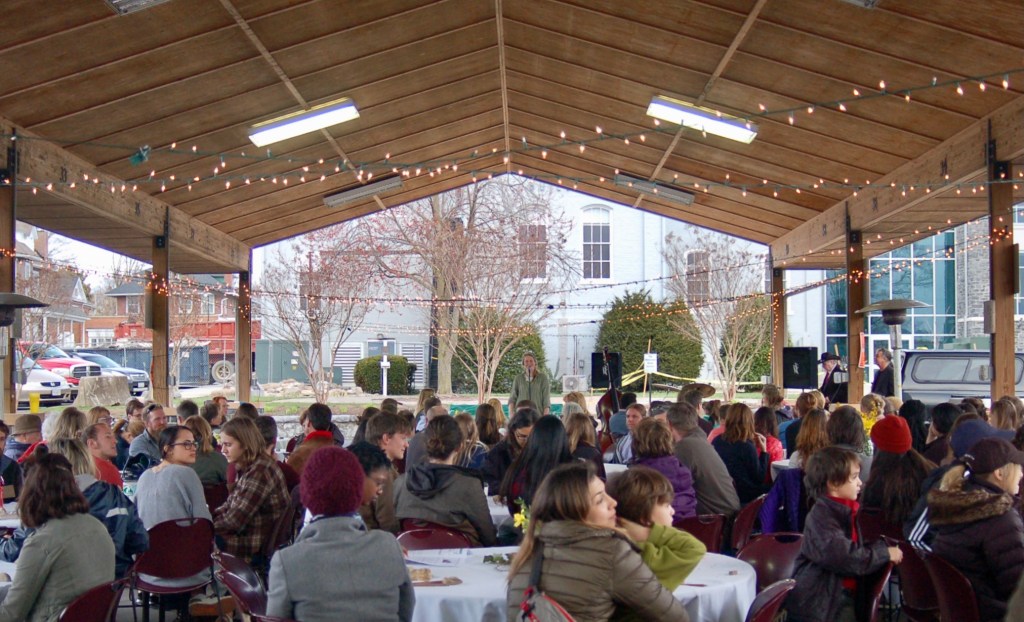 Livvy stands at the far end of the wood paneled pavilion to welcome the people sitting around tables. String lights hang above the crowd. 