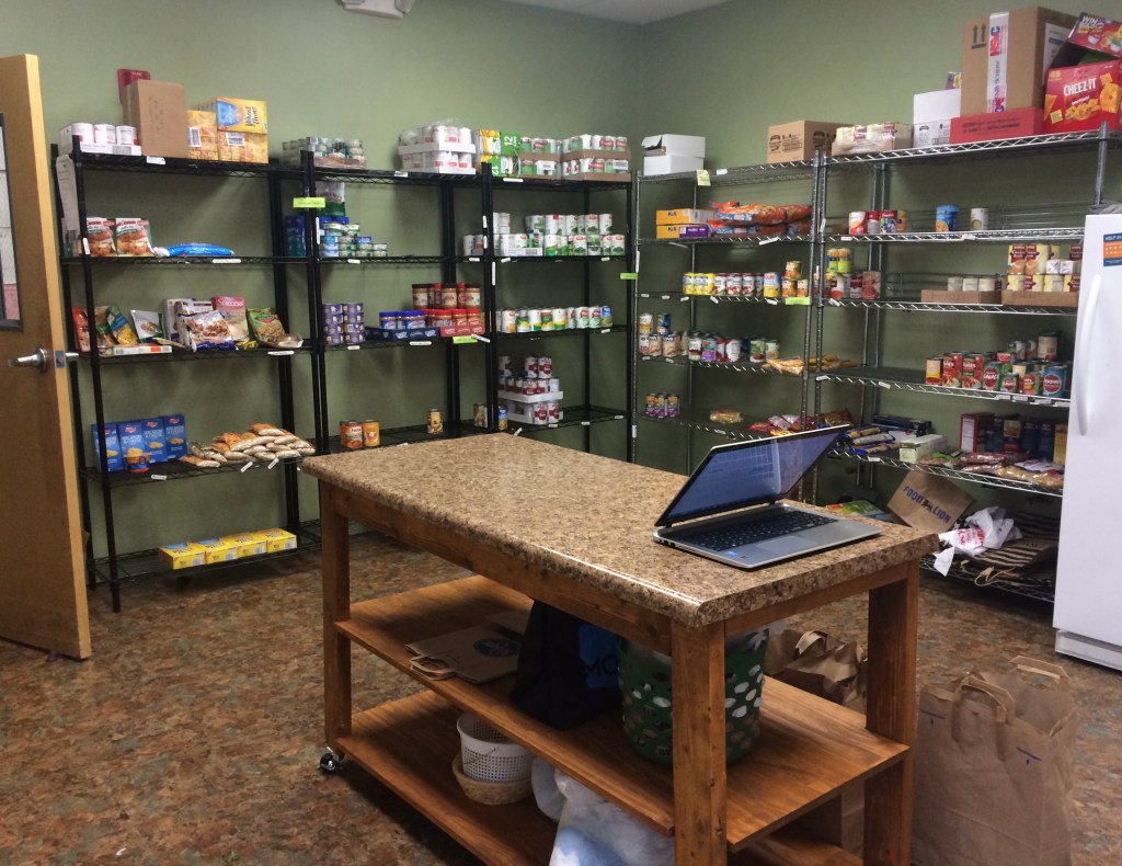 A room with tall shelves that line the wall next to a refrigerator. Food cans and boxes sit on the shelves. An island sits in the middle of the room with a computer on top.
